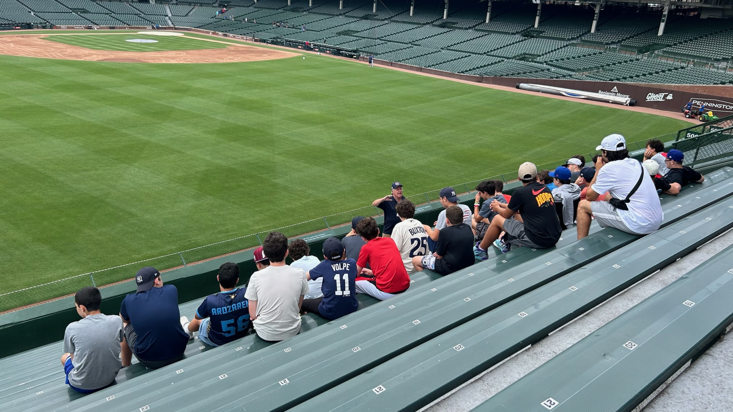 Group of people sitting on green stadium bleachers overlooking a baseball field in a large ballpark.