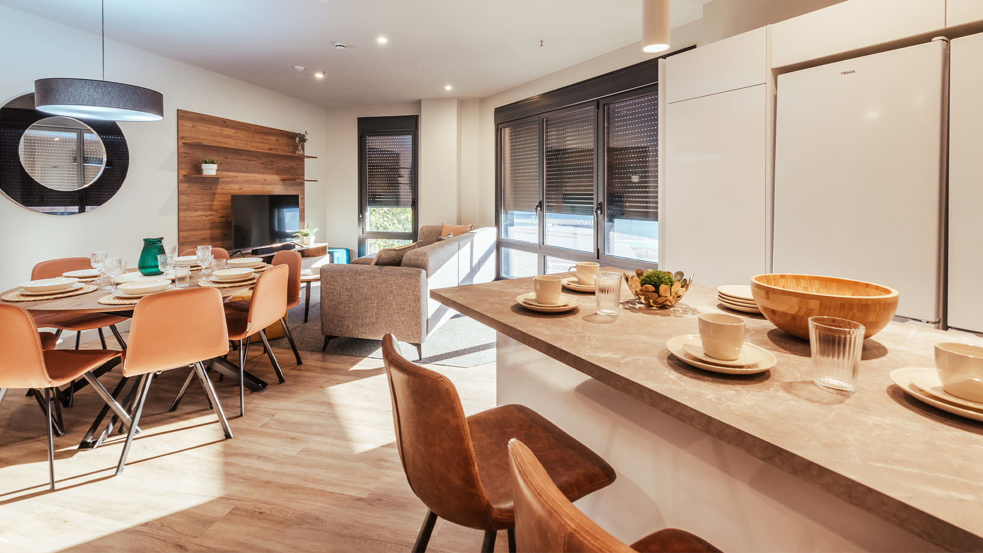 Open-plan dining and living area with a round table set for eight, orange chairs, and a marble kitchen island bathed in sunlight from large windows.