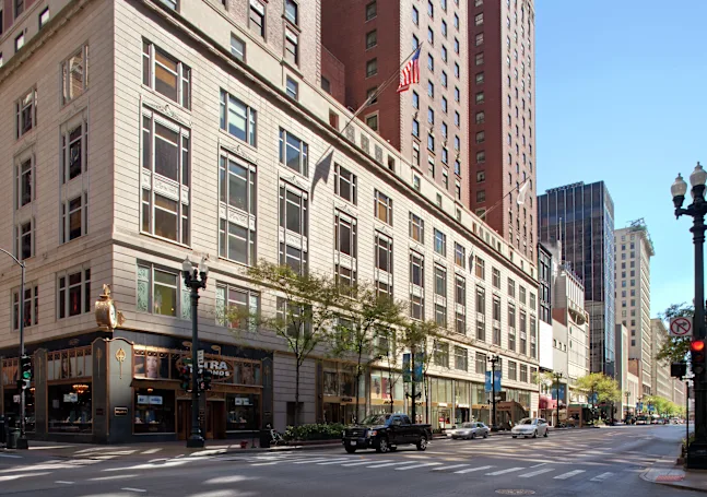 City street scene with a beige multi-story building and storefronts along the curb, American flag overhead.