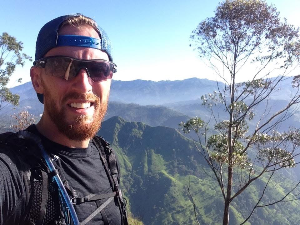 Hiker wearing a blue backward cap and sunglasses on a mountain ridge overlook with trees and distant valleys under a clear sky.