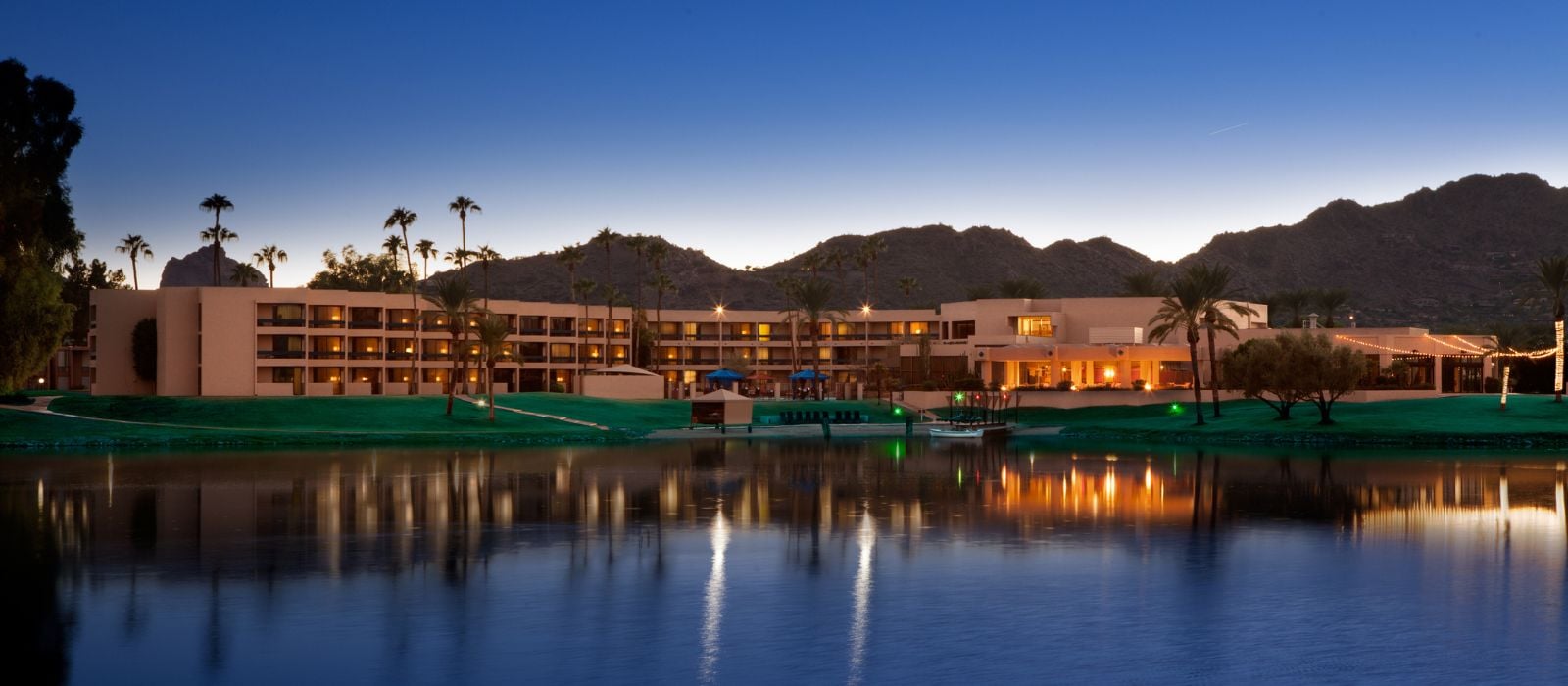 Resort hotel beside a reflective lake at dusk, with palm trees and mountains in the background and warm lights.