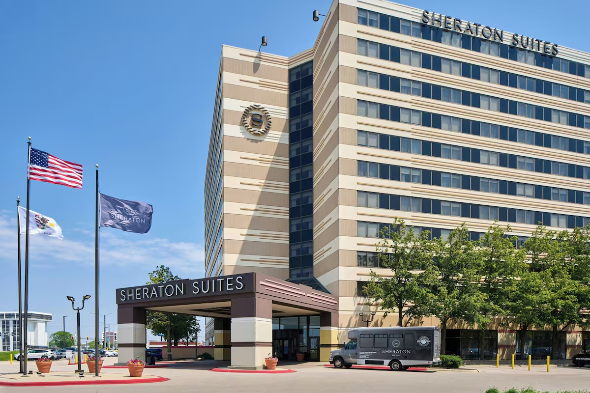 Front view of a Sheraton Suites hotel with a tall beige tower, entrance canopy, and flags in the foreground.