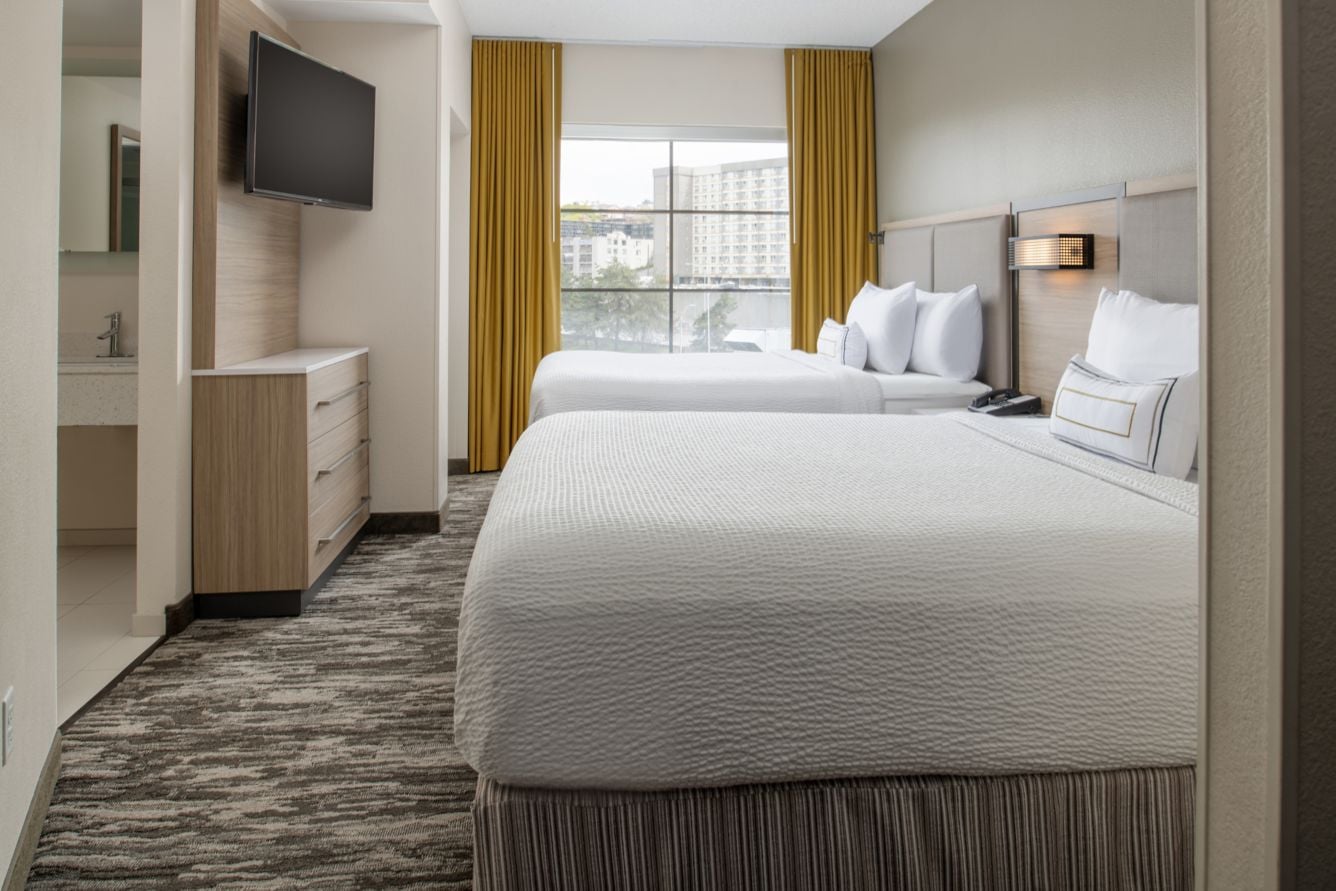 Hotel room with two queen beds, white bedding, and a large window with mustard-yellow curtains; wall-mounted TV above a dresser.