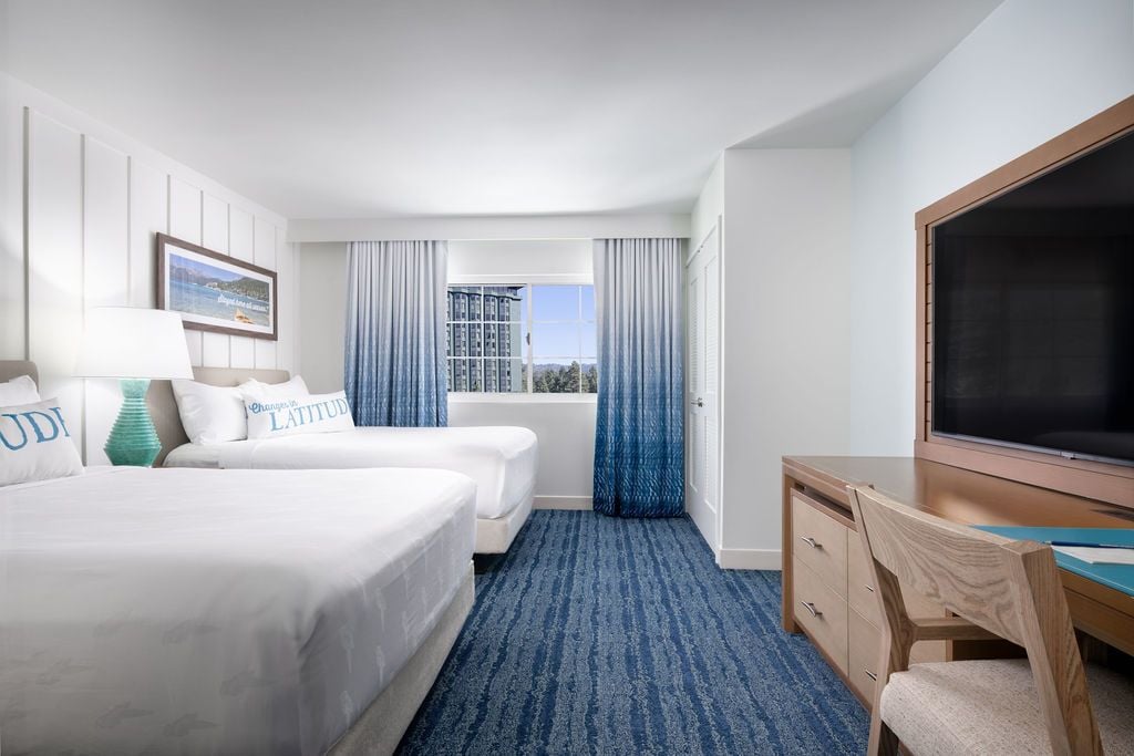 Hotel room with two white beds, turquoise lamp, framed landscape photo, blue gradient curtains, and a desk with a flat-screen TV.