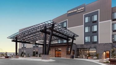 Exterior of a SpringHill Suites by Marriott hotel with a glass-and-metal porte-cochère and parking area beneath a clear blue sky.