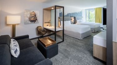 Modern hotel suite with a living area, glass partition, and two queen beds by a large window in a bright room.