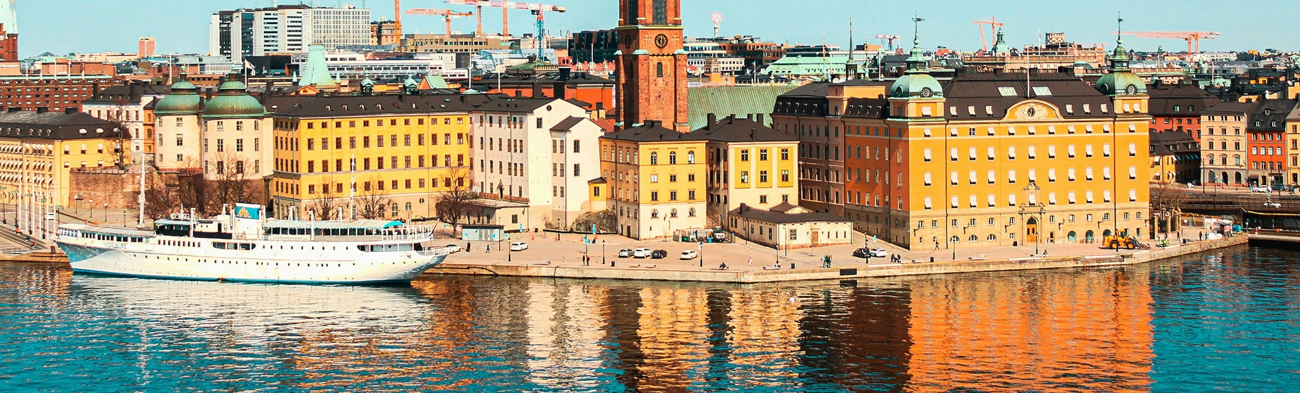Colorful historic harbor with yellow, orange buildings along a quay and a white passenger ship docked by the water, reflections shimmering on the surface